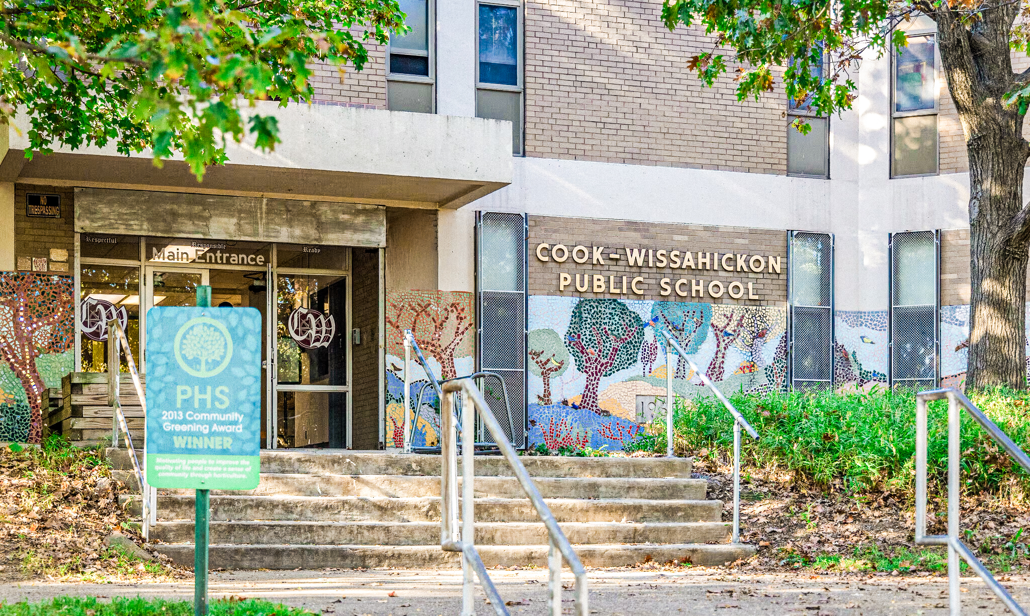Cook-Wissahickon Public School Building The front entrance of Cook-Wissahickon Public School, featuring a colorful tree mosaic along the lower walls, a “Main Entrance” sign above the doors, and a PHS 2013 Community Greening Award sign in the foreground.